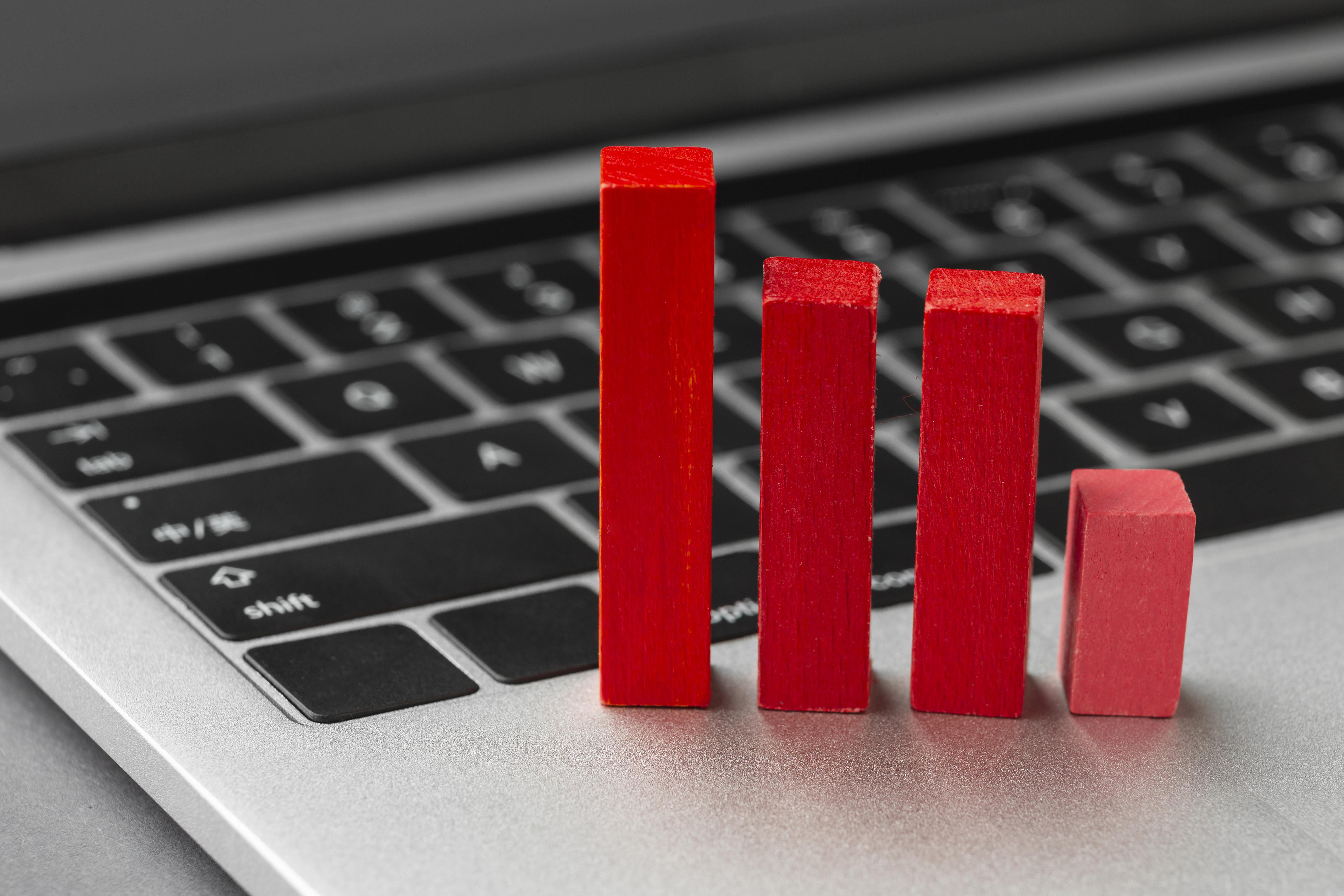 A close-up of a laptop keyboard with four red wooden blocks of varying heights placed on it, representing a bar chart or declining statistics. The background is blurred, and the image conveys a concept related to business, analytics, or digital marketing trends.